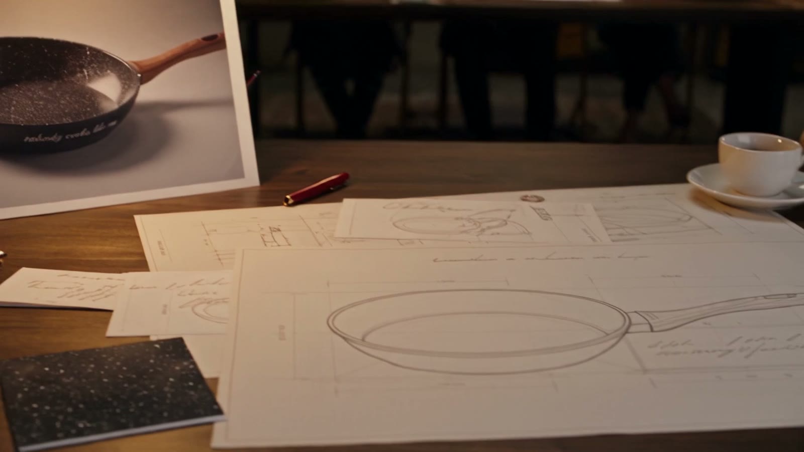 Una riunione attorno a un tavolo, con disegni tecnici e la fotografia di una padella appoggiata sul piano